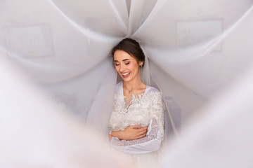 Wedding portait of happy bride in white wedding veil. Gorgeous beauty young bride portrait. Beautiful bride with wedding makeup holding wedding dress. Bridal fashion model posing in interior.