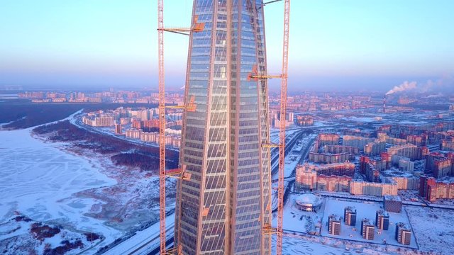 Glass walled twisting facade of supertall skyscraper, construction of tallest building in Europe in progress, tower cranes attached from both sides Lakhta Center. Aerial shot against city quarters