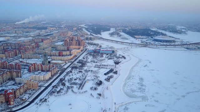 Aerial shot of large coastal park and Primorsky District of Saint Petersburg. Winter evening time, frozen Neva bay water on right side, dim cityscape and horizon in mist, stadium and highway road