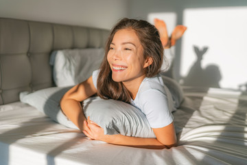 Bed woman waking up early morning happy enjoying sun on comfortable mattress and pillow. Asian girl relaxing in bedroom smiling. Natural beauty after healthy good night sleep sleeping in on weekend.