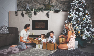 Christmas mood. Family of mom, dad, daughter and son open their gifts. Decorated Christmas tree and fireplace on the background. Interior in grey color
