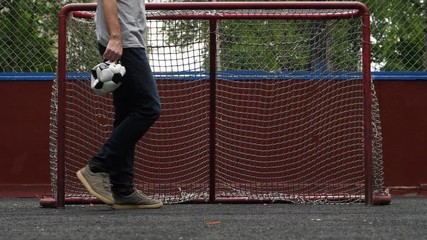 Man walk with deflated soccer ball in hand against small gate, slow motion shot. Player pass by, dangle hand with broken ball.