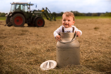 An adorably Bavarian baby boy standing in a milk churn laughing happily. In the background a tractor is turning  the hay the milk churn in standing on.