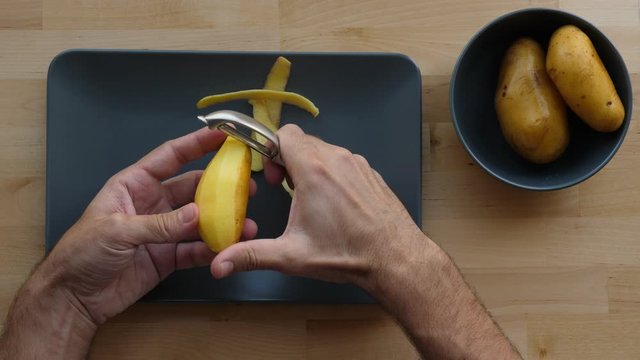 Man clean yellow potato using special tool, top-down shot. Cook prepare basic ingredient, remove skin from popular vegetable, peelings fall down on rectangle plate