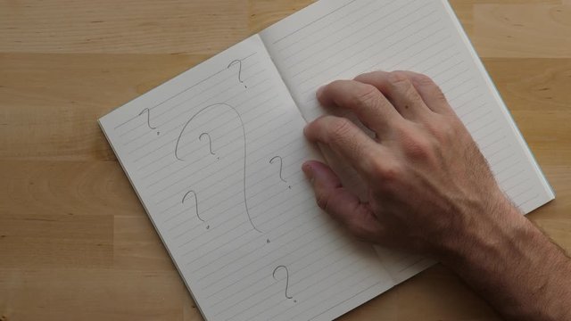 Man think over notepad with only question signs written on page, tap fingers, top down shot of wooden desktop. Hard problem or too many questions