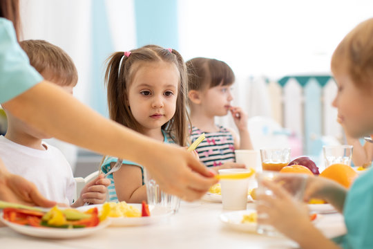 Little boys and girls from the group of preschool children sit at table with lunch and eat appetizing in kindergarten. Kids with caregiver in day care centre