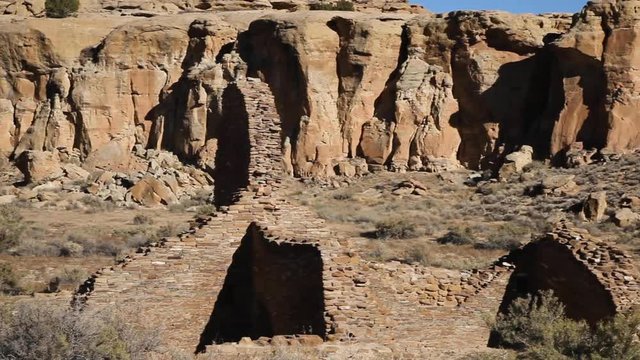 Panning across Anasazi ruins at Chetro Ketl, a religious site used thousands of years ago in Chaco Canyon, New Mexico. The brick buildings blend in with the rocky desert cliffs behind them. Shadows.