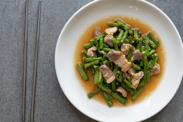 Fried yardlong beans with streaky pork in white dish on concrete table.