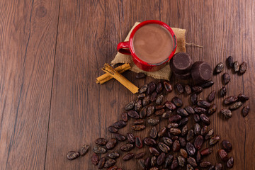Red cup with hot chocolate on wood surrounded by cocoa beans, pieces of pure chocolate and cinnamon - pewter cup top view