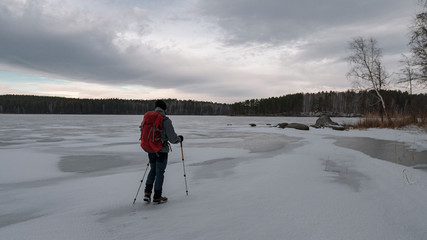 Young man walking on frozen lake with backpack and trekking poles; cloudy dramatic winter sky, cold...