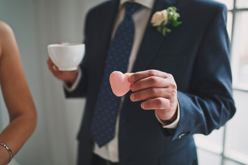 Crop groom is holding macaroon in the form of a pink heart and drinking tea or coffee.