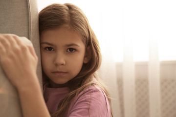 Sad little girl hiding behind sofa at home. Domestic violence concept