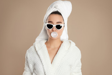 Young woman in bathrobe and sunglasses blowing chewing gum on brown background