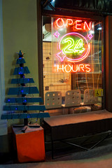 Open 24 hours neon sign on cafe, wooden Christmas tree with garlands at night in winter