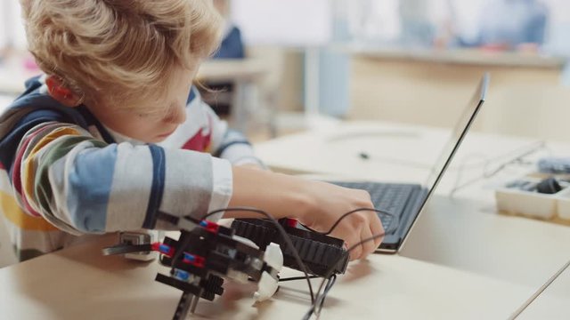 Schoolboy Connects Cables to Small Robot, Uses Laptop to Program Software for Robotics Engineering Class. Elementary School Science Classroom with Gifted Brilliant Children Working with Technology
