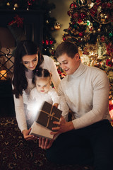 Three members of family opening Christmas gift together. Mother, father and little daughter opening magic present for Christmas with illumination inside. Family is sitting in front of the beautiful