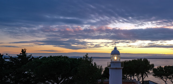 A working Lighthouse on romantic, dramatic autumn sunset over the calm sea background. Fantastic colors of the sky. Natural gradient. The concept of safe sea navigation. Roses, Catalonia, Spain.