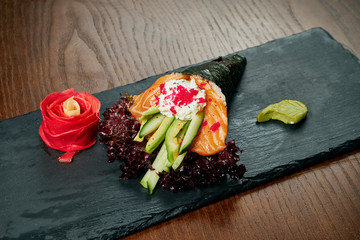 Close-up of tasty hand roll sushi in mamenori with salmon and Tobico caviar served on dark stone plate with soy sauce and ginger. Copy space. Temaki, Japanese cuisine. Healthy food