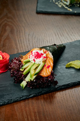 Close-up of tasty hand roll sushi in mamenori with salmon and Tobico caviar served on dark stone plate with soy sauce and ginger. Copy space. Temaki, Japanese cuisine. Healthy fooda
