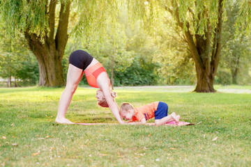 Family parental activity. Young Caucasian mother with child toddler boy doing workout yoga fitness outdoor in park on summer day. Woman doing sport exercises together with her kid.