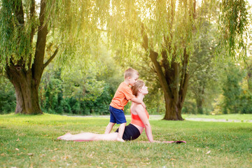 Family parental activity. Young Caucasian mother with child toddler boy doing workout yoga fitness outdoor in park on summer day. Woman doing sport exercises together with her kid.
