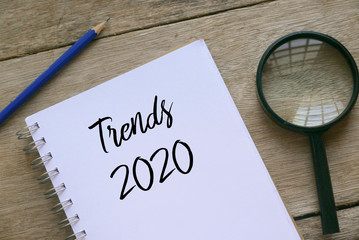 Top view of magnifying glass,pencil and notebook written with Trends 2020 on wooden background.