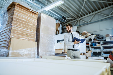 Fototapeta premium Smiling caucasian bearded supervisor in tie and suit looking at pile of sheets and writing down inventory. Printing shop interior.