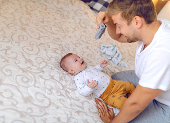 Happy handsome caucasian young dad holding tiny baby socks and preparing to change clothes to his laughing cute 6 months old son. Bedroom interior.