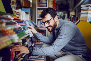 Young handsome caucasian bearded man with eyeglasses crouching in bookstore and searching for...