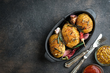 Baked potatoes with herbs, spices, garlic and sauce in a black pan on a dark background top view free copy space.