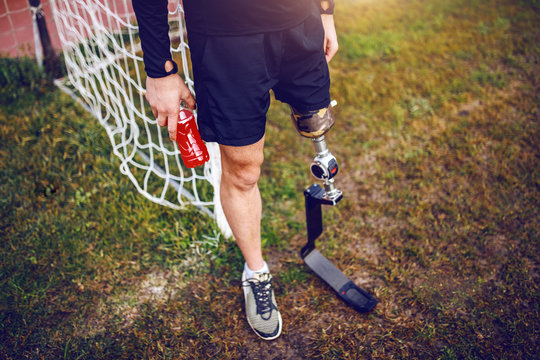 Handsome sporty handicapped man in sportswear and with artificial leg while standing on football court and holding refreshment. - Powered by Adobe