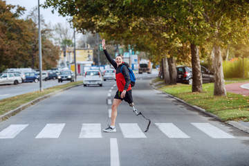 Full length of handsome caucasian handicapped sportsman in sportswear, with artificial leg and backpack crossing street and waving to a friend.