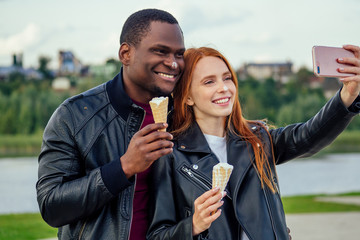 Multi ethnic diverse couple eating ice cream in spring autumn park and making selfie photo on camera of smartphone