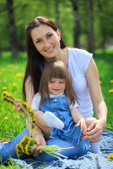 Happy smiling  mother and her little daughter weaving a wreath of dandelions in the park