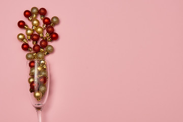 Champagne glass with golden and red baubles on pastel pink background, copy space. Minimal, New year concept. Top view, flat lay