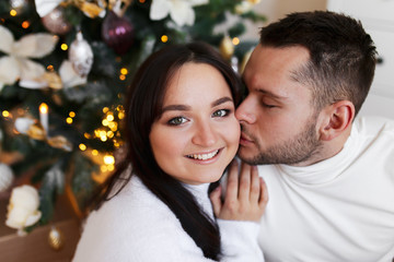 Fototapeta premium An attractive brunette girl in a white wool dress and a brutal man in a sweater hug and kiss on the background of a Christmas tree, a chest of drawers and decorative stars. happy family and Christmas.