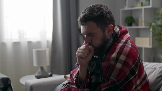 A man coughs heavily while sitting in a plaid on a couch in the living room