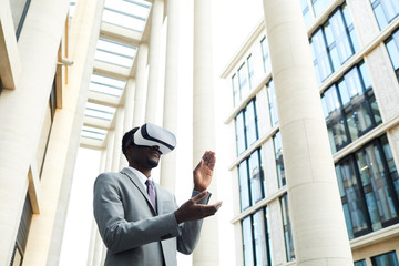 Serious African businessman in suit wearing goggles and clapping while playing in virtual reality game outdoors