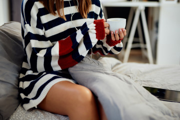 Caucasian woman in striped sweater sitting on bed, listening music over tablet and holding her fresh morning coffee.