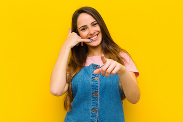 young pretty woman smiling cheerfully and pointing to camera while making a call you later gesture,...
