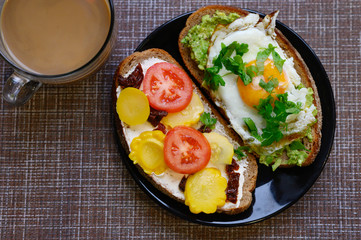 breakfast and brunch. two sandwiches of black bread, with cheese and vegetable filling, lie in a black plate, next to a cup of coffee with milk on a textile background