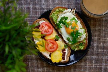 breakfast and brunch. two sandwiches of black bread, with cheese and vegetable filling, lie in a black plate, next to a cup of coffee with milk and a green plant in a white pot on a textile background