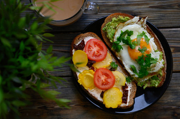 breakfast and brunch. two sandwiches of black bread, with cheese and vegetable filling, lie in a black plate, next to a cup of coffee with milk and a green plant on a wooden background