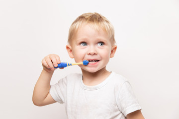 little cute caucasian blond blue-eyed boy in a white t-shirt brushes his teeth on a white background