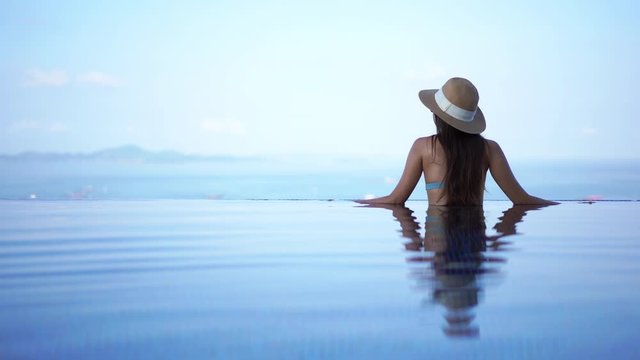 Water ripples around a young woman standing in the shallow water at the edge of a hotel pool looking out at the seascape,