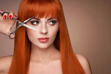 Fototapeten Beautiful young woman with a bright makeup and a smooth long hair holds metal scissors. Model with red hair. Hair salon, haircut. Care and beauty hair products. Perfect make-up © Oleg Gekman