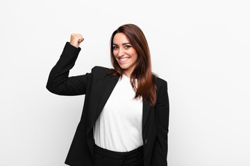 young pretty businesswoman feeling happy, satisfied and powerful, flexing fit and muscular biceps, looking strong after the gym against white wall