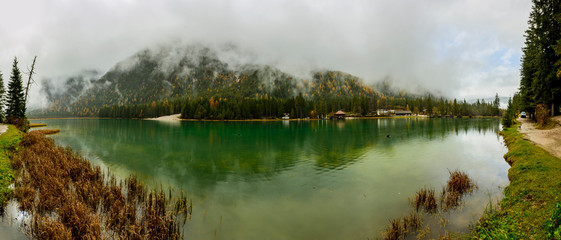 Fototapeta na wymiar Boat houses in turquoise colour lake South Tyrol, Italy , Alps. Reflection in water during rain (high ISO long exposure image) Dark green forest in background.