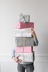 In the man&Otilde;s hands is a huge stack of packed holiday boxes, gray and pink color. Gifts for the new year, Christmas, birthday or other holiday. Against the background of a white wall.
