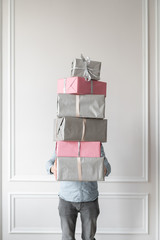 In the manÕs hands is a huge stack of packed holiday boxes, gray and pink color. Gifts for the new year, Christmas, birthday or other holiday. Against the background of a white wall.